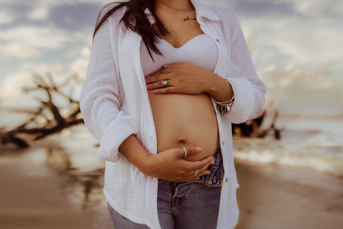 Pregnant person standing on a beach, wearing a white top and holding their belly with both hands, with driftwood and waves in the background.