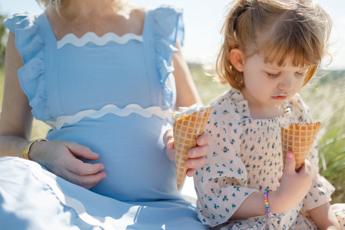 a pregnant mother and her daughter share ice cream cones during a maternity photoshoot on a hillside in the warm summer day
