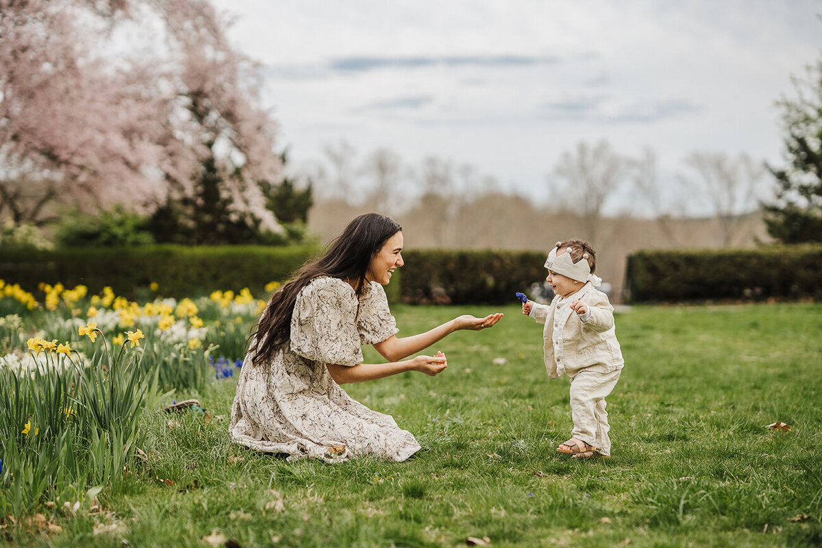 mother holds arms out to baby boy taking first steps during boston family photo session