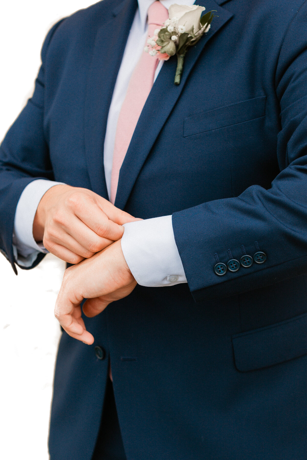 Groom adjusting his cufflinks in a navy suit and pink tie. A stylish detail shot focusing on the groom's attire, showcasing his preparation for the big day.