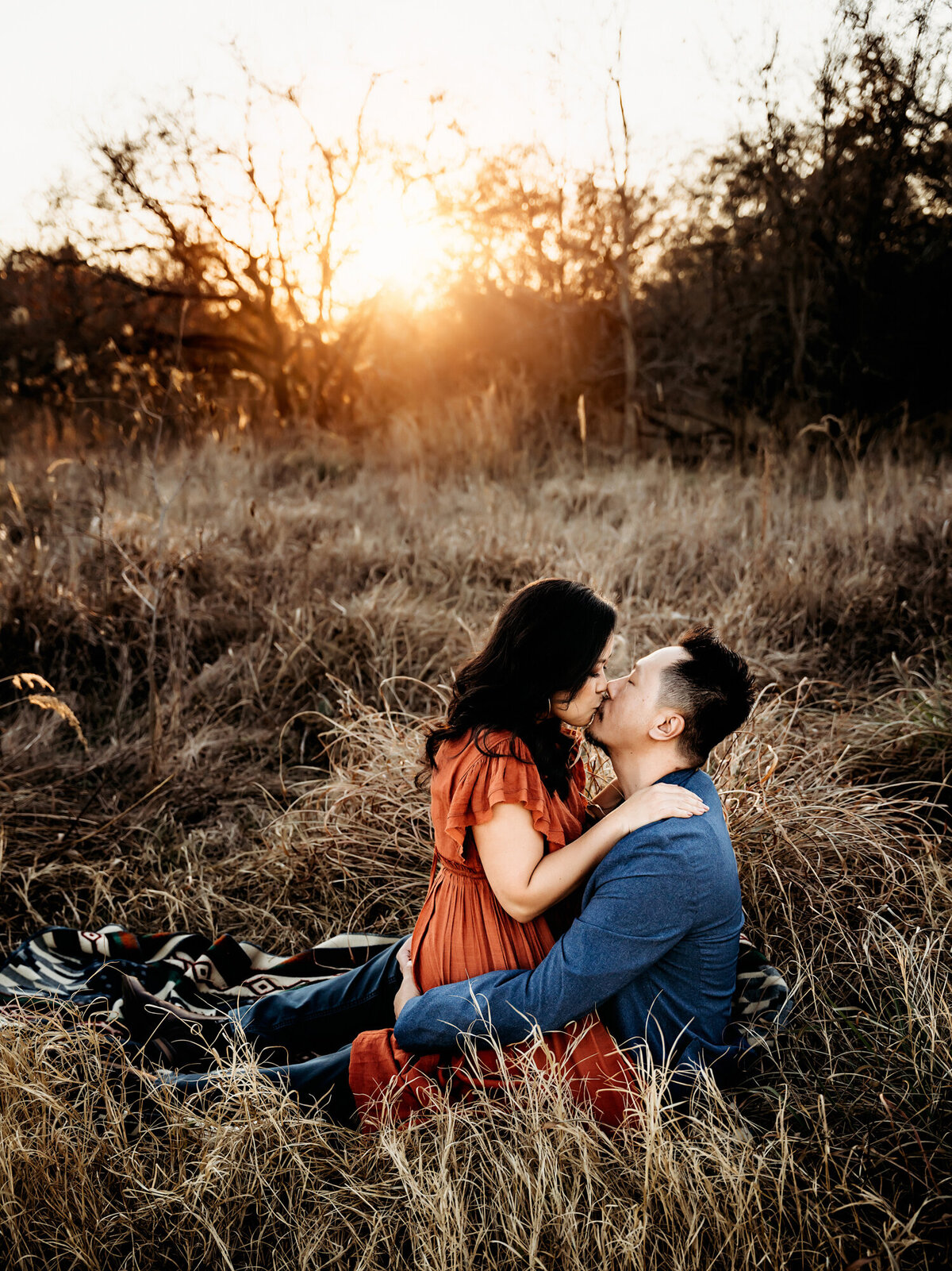 Couples Photography, Woman in a rust dress straddling man in a blue suit and kissing him, in the field at sunset on a blanket.