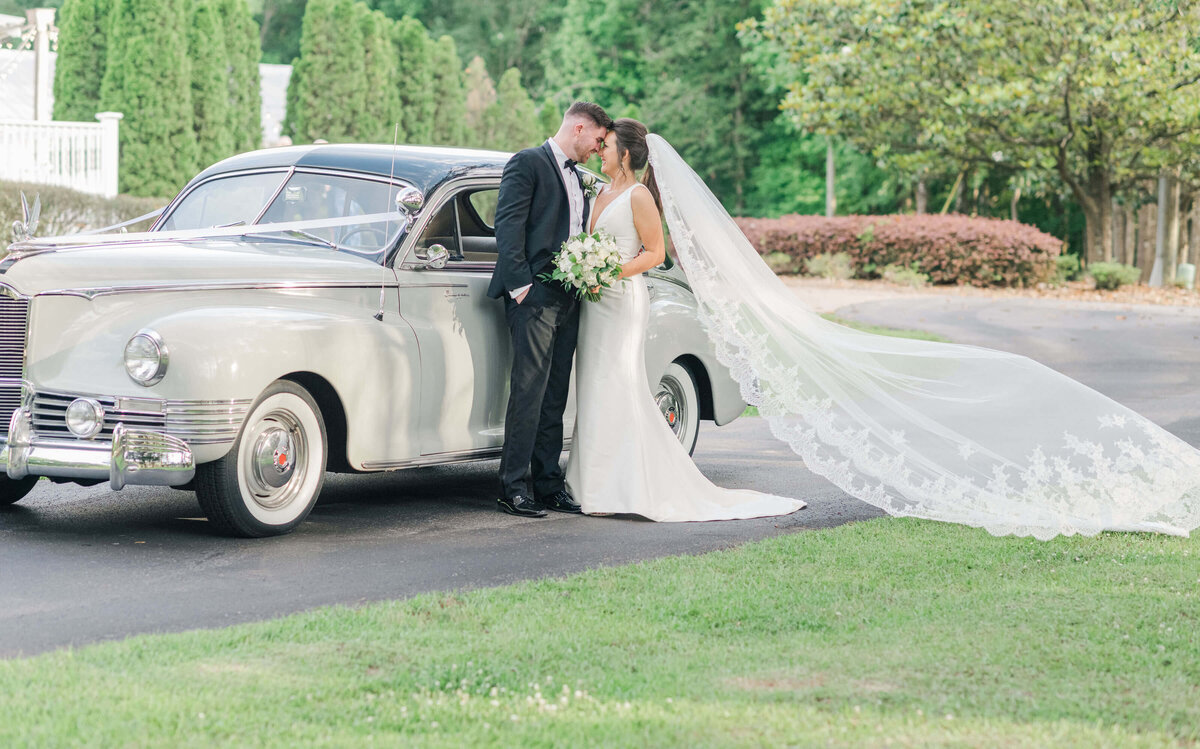 Bride and groom looking at each other while leaning against a classic car with brides veil flowing