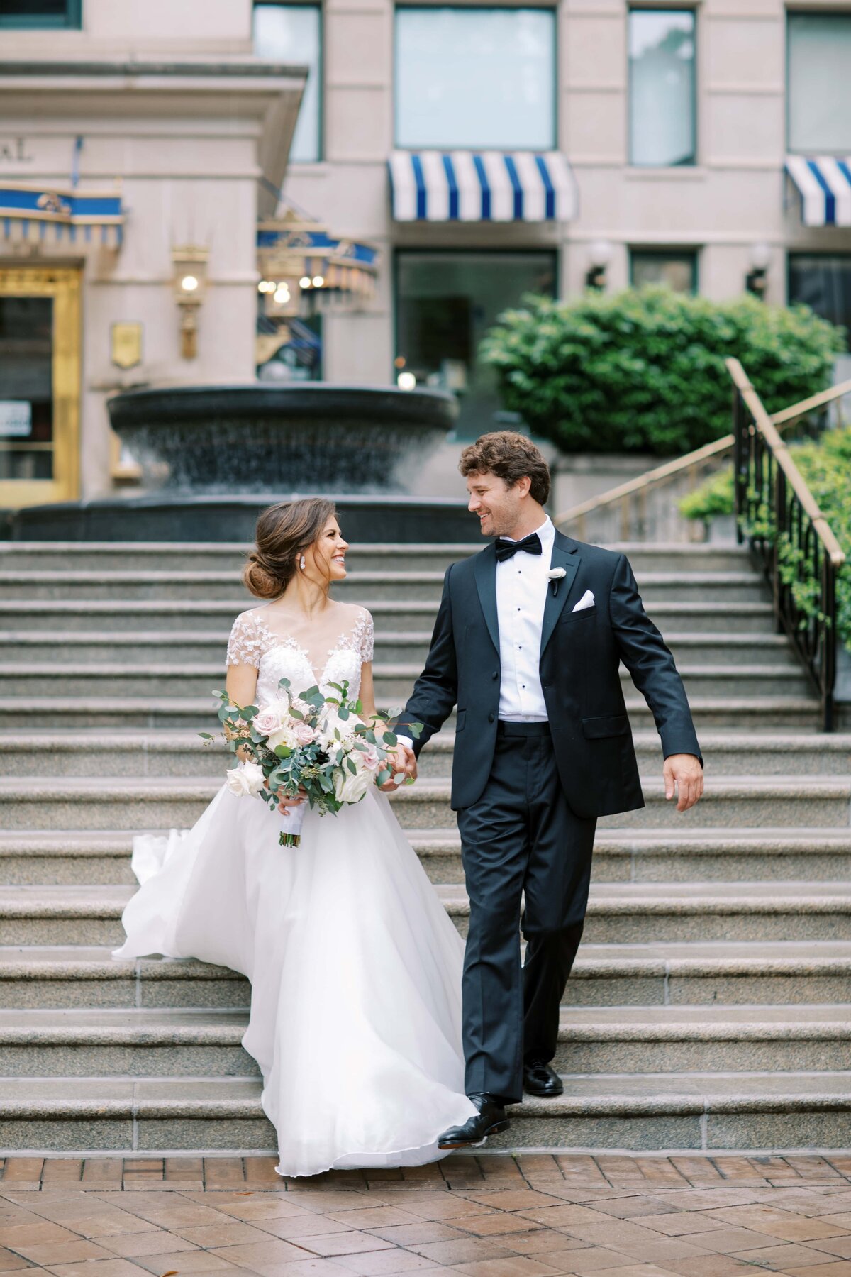 Stunning young bride with brown hair and in wedding dress holding the hand of a handsome groom in a black tux  as they smile at each other and walk down the outdoor staircase at The Willard Hotel in Washington DC