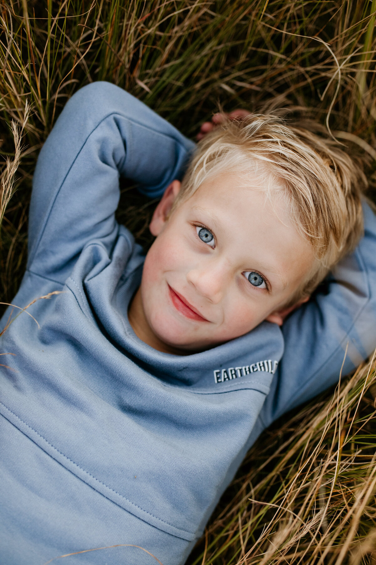 A young boy with icy blue eyes is lying in a field, with his arms behind his ears, looking relaxed.
