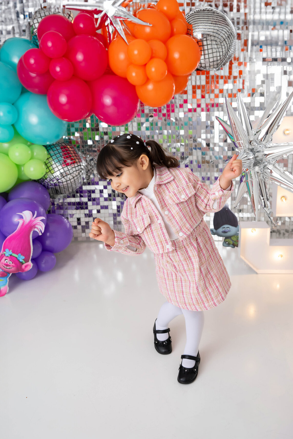 Child stands in a modeling pose by a shimmering backdrop surrounded by colorful balloons.