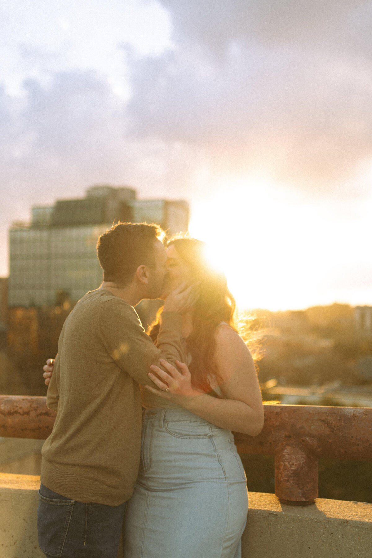 a sunset photo of a couple kissing on top of a parking garage and the sunlight is beaming behind them.