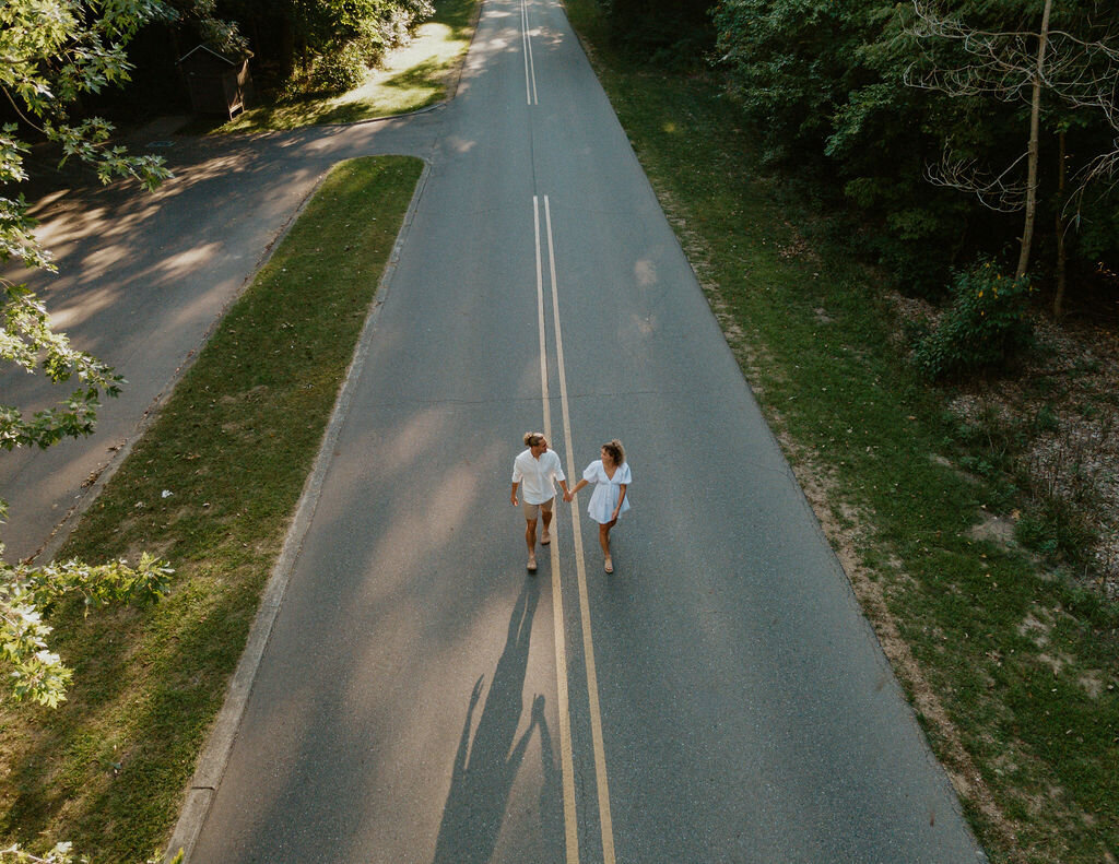 Arial view of couple wearing white holding hands, looking at each other, and walking down a two lane road