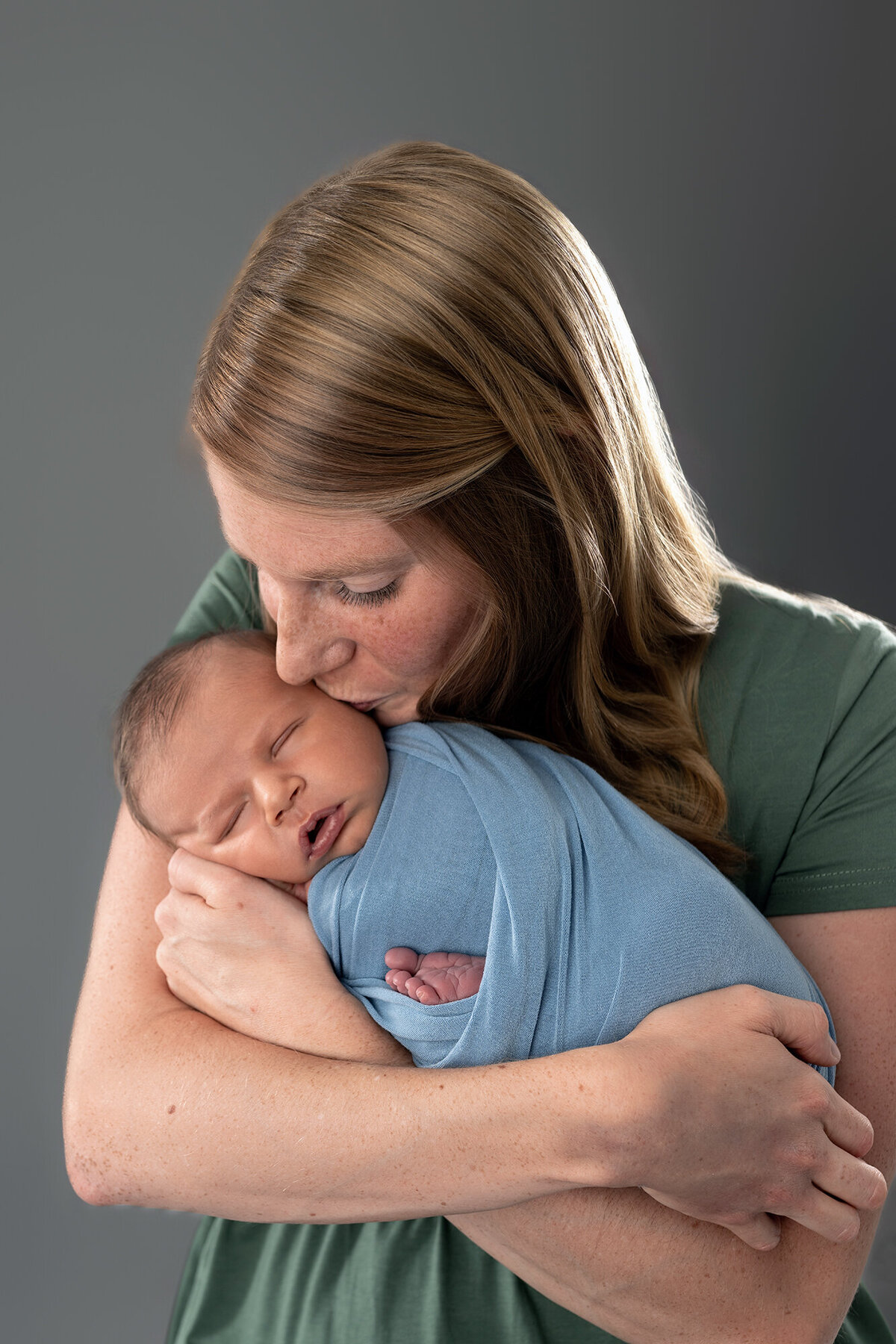 A happy mom kisses her newborn son in her arms while standing in the studio of a Des Moines Newborn Photographer
