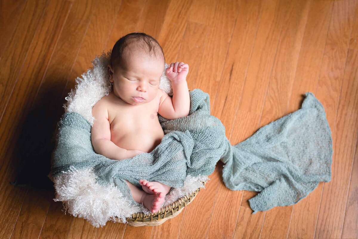 Newborn baby asleep in a basket, loosely wrapped in a teal baby wrap in Columbia SC