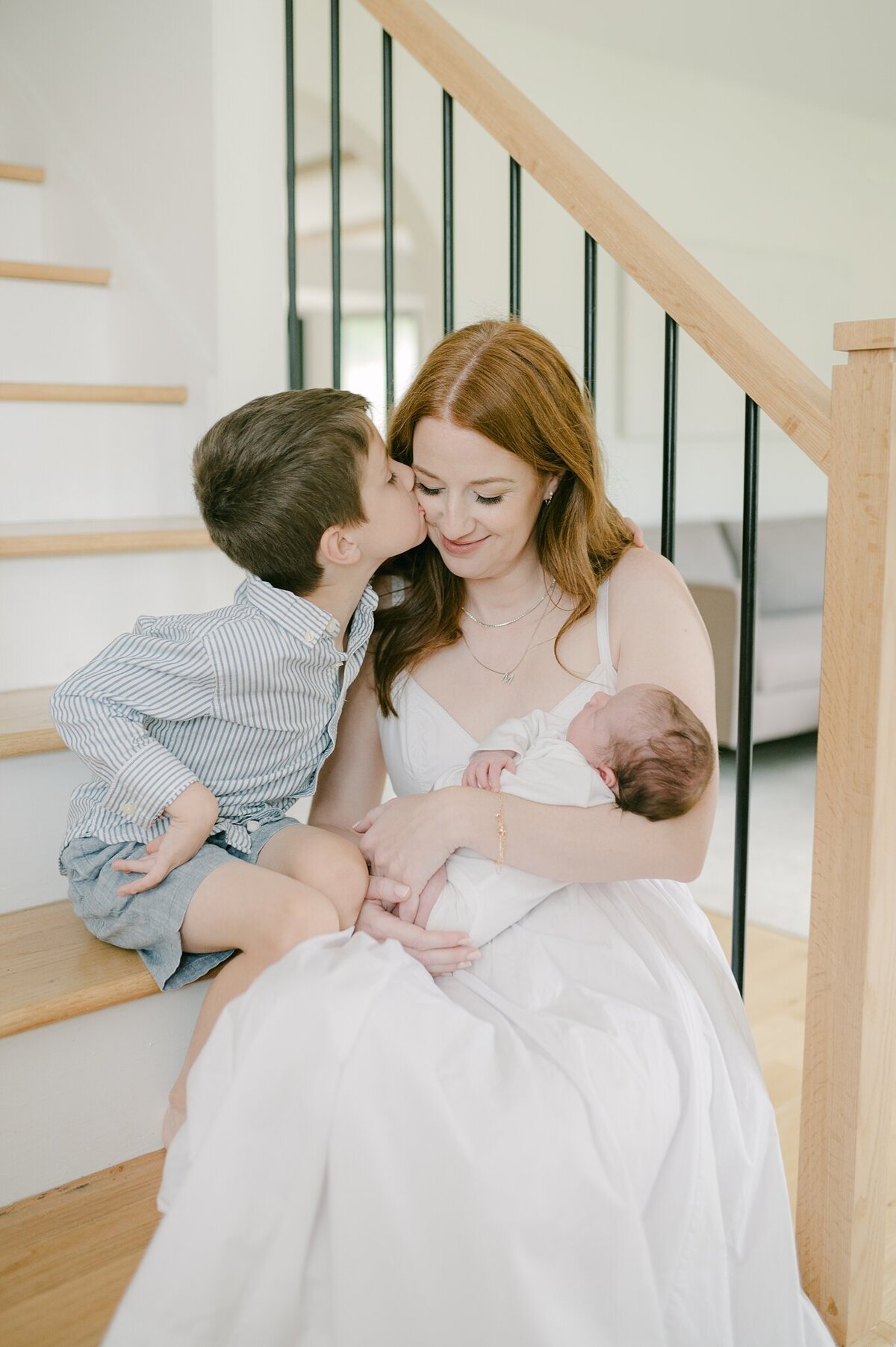 Mom is sitting on her staircase holding her newborn baby and her son is next to her holding her cheek. She is smiling down.