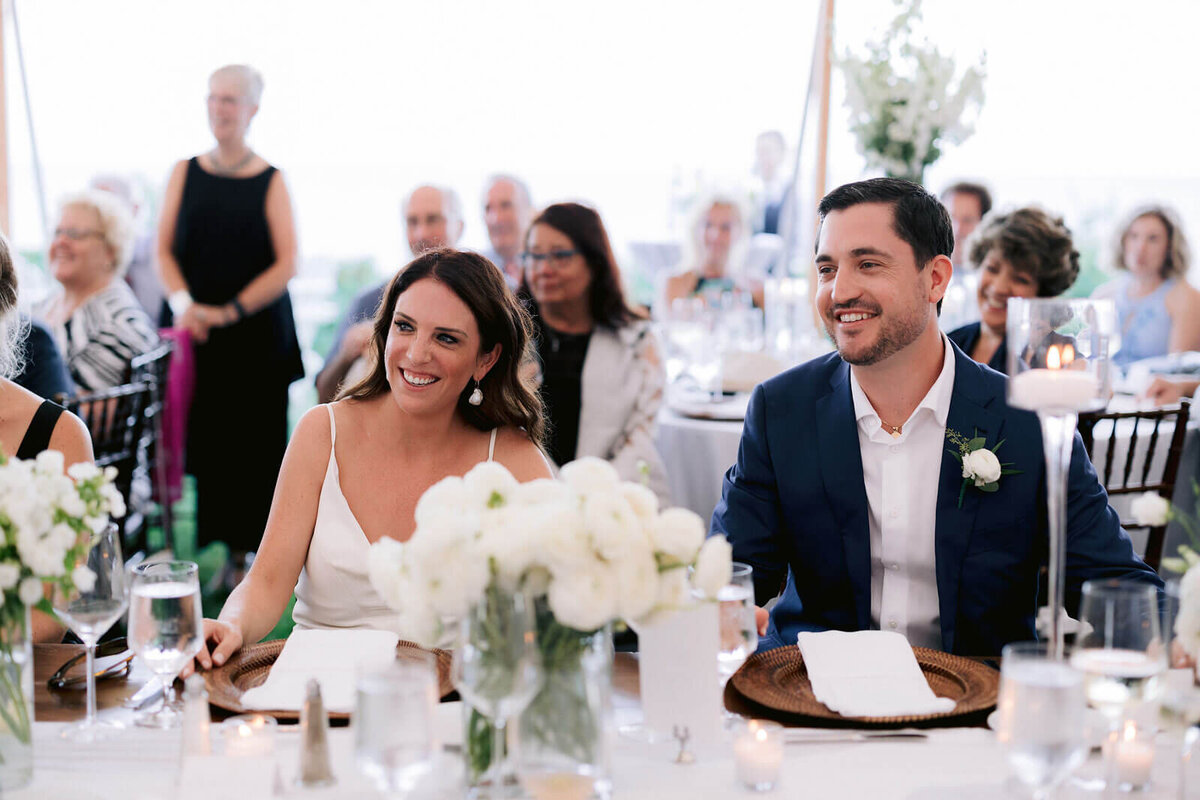 The bride and the groom are smiling while sitting at the dining table, guests in the background, at Cape Cod Summer Tent, MA.