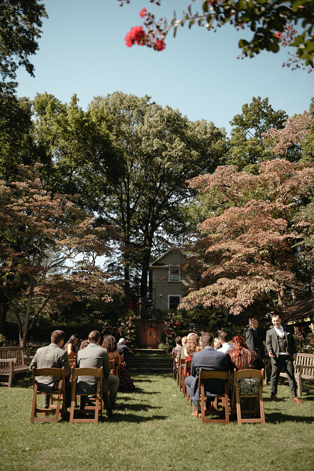 An intimate garden wedding ceremony with guests seated on wooden chairs, surrounded by lush greenery and warm autumn foliage.