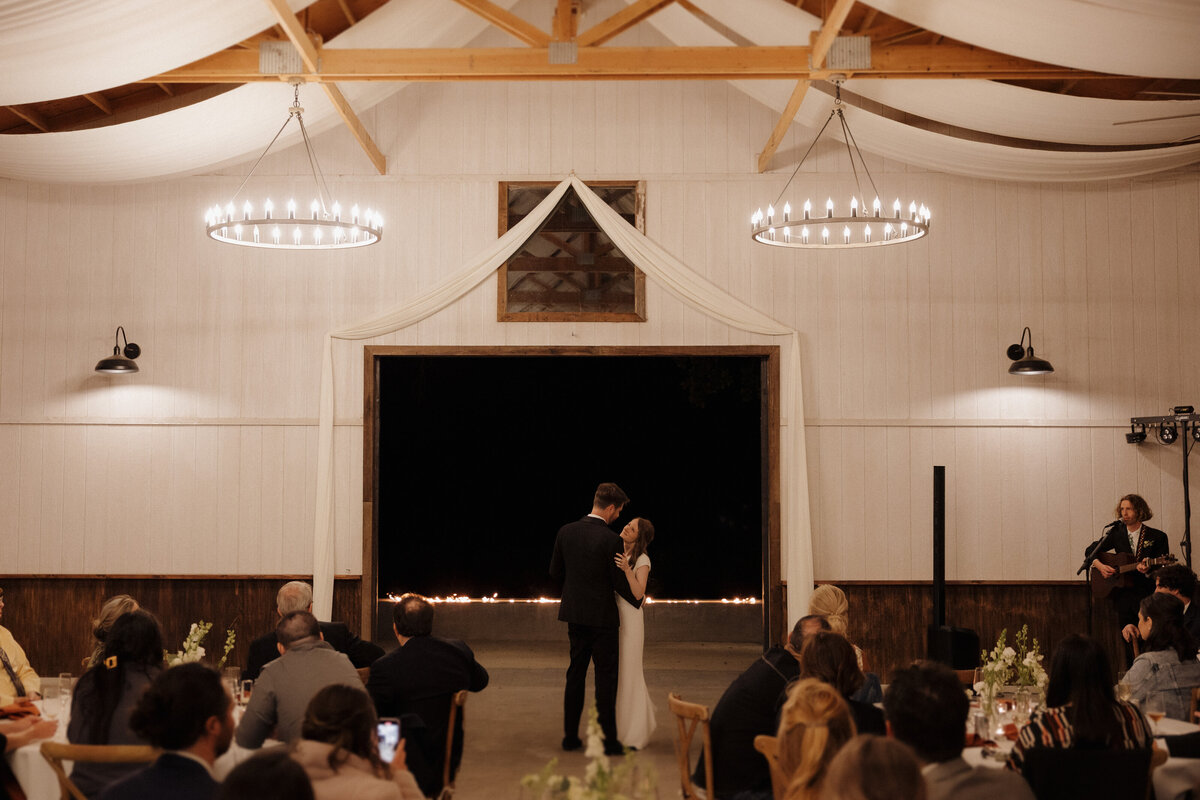 A bride and groom share a romantic first dance in a candlelit barn with an open-air backdrop, while a live musician serenades them.