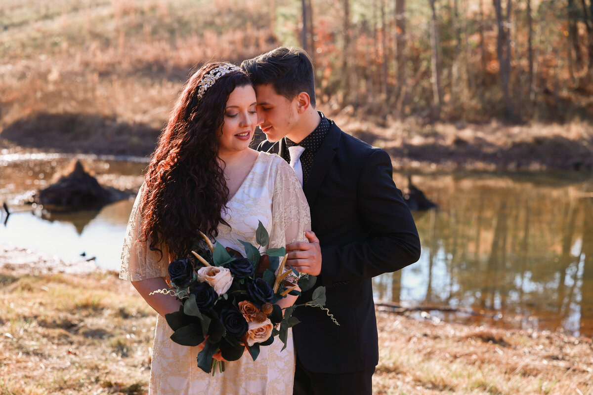 bride holding blue and maroon floral bouquet with groom standing behind her in front of a pond in Rome Georgia