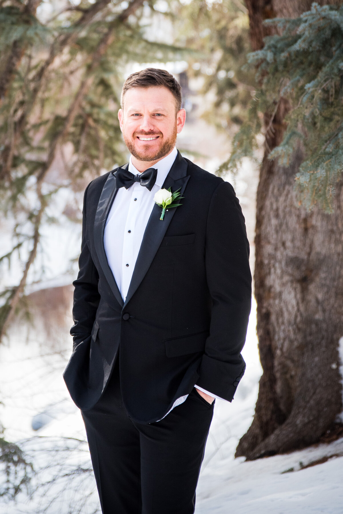 A groom smiles at the camera with his hands in his pockets in a snowy setting in Vail, Colorado.