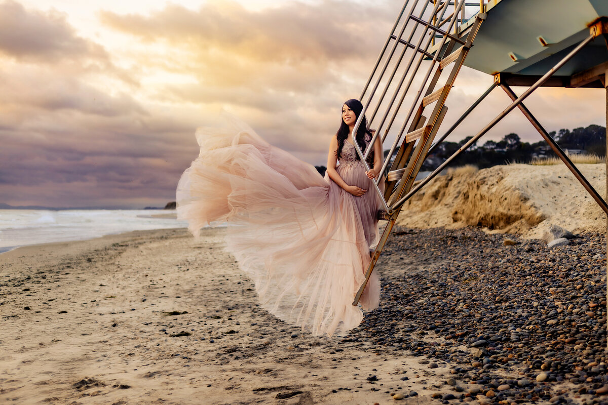 San Diego maternity photographer, a mom stands on a lifeguard tower at the beach in a maternity gown.
