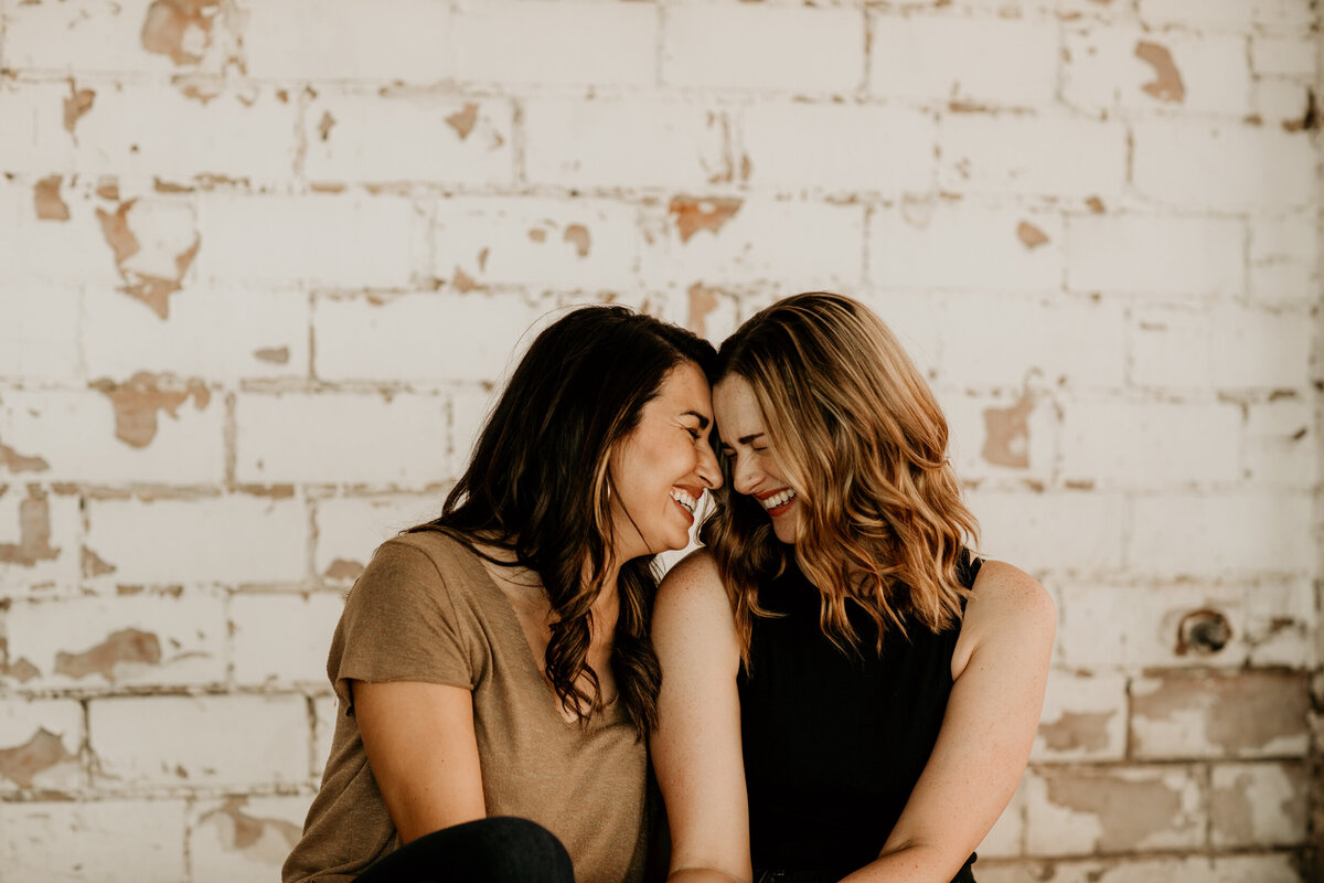 same sex couple laughing  and sitting together in front of a white brick wall