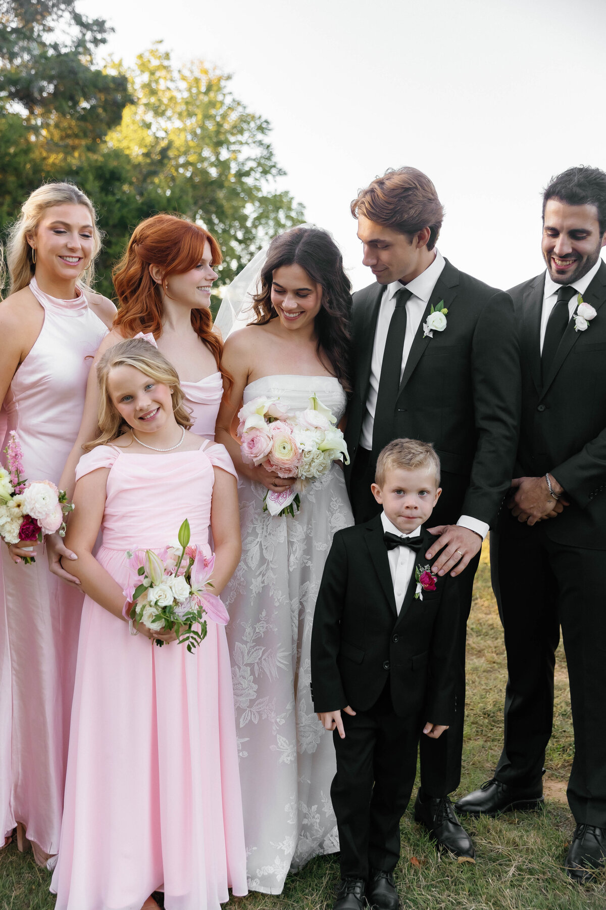 Bride and groom pose with their wedding party, including bridesmaids in blush dresses and children holding petite floral bouquets styled by a fine-art wedding florist in Arkansas.