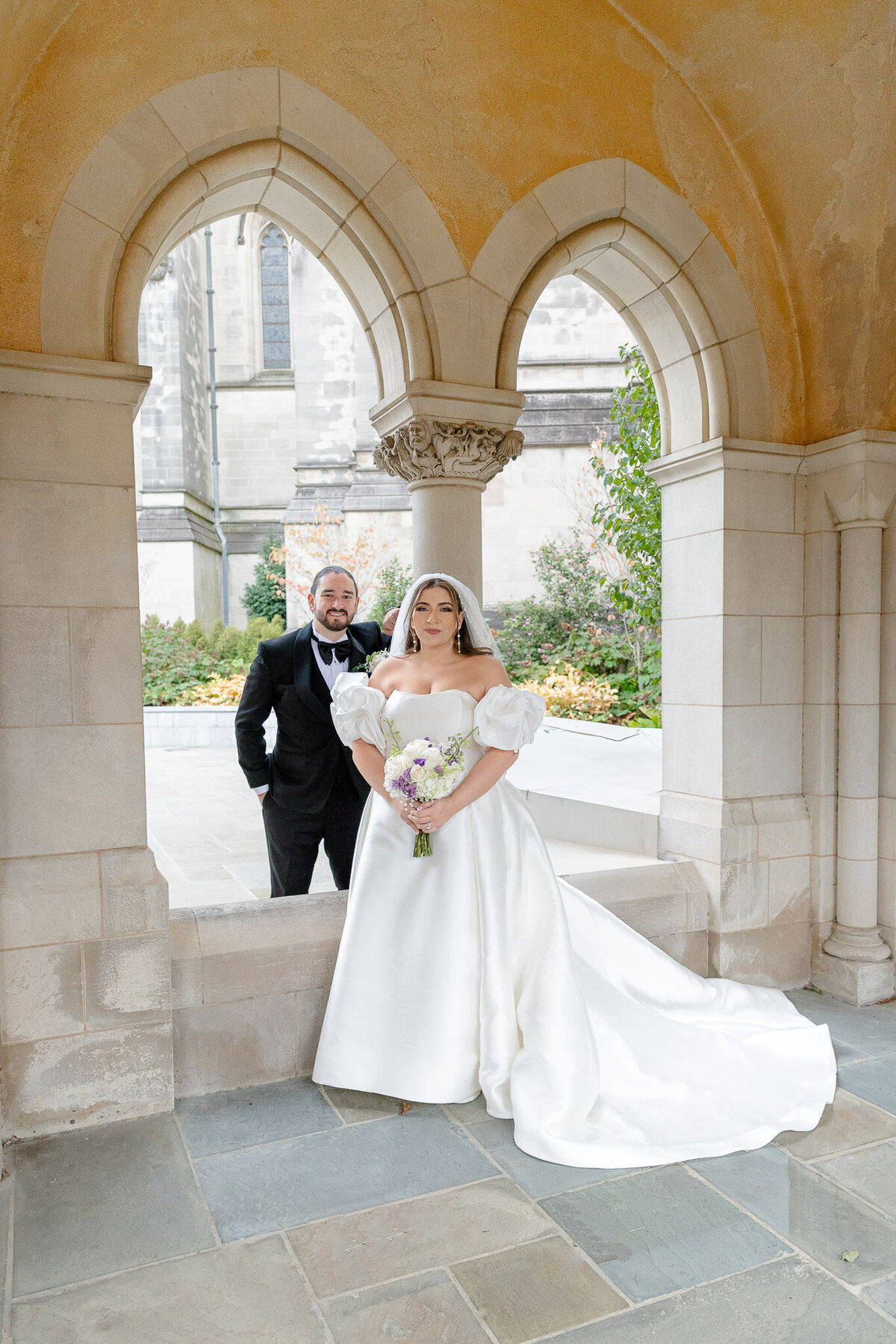 A bride and groom pose in a stunning cathedral under pointed arches. They are both softly smiling at the camera.