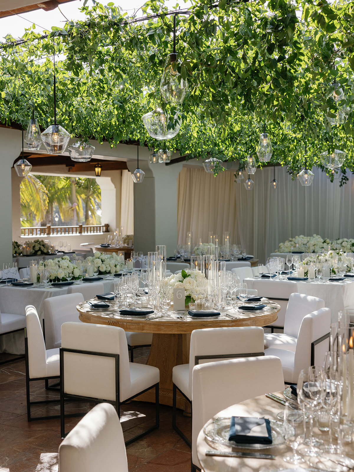 Wedding reception at The Ritz Carlton Bacara with in outdoor courtyard with greenery overhead on lighting, white upholstered dining chairs, white draping, white flowers, and wood tables