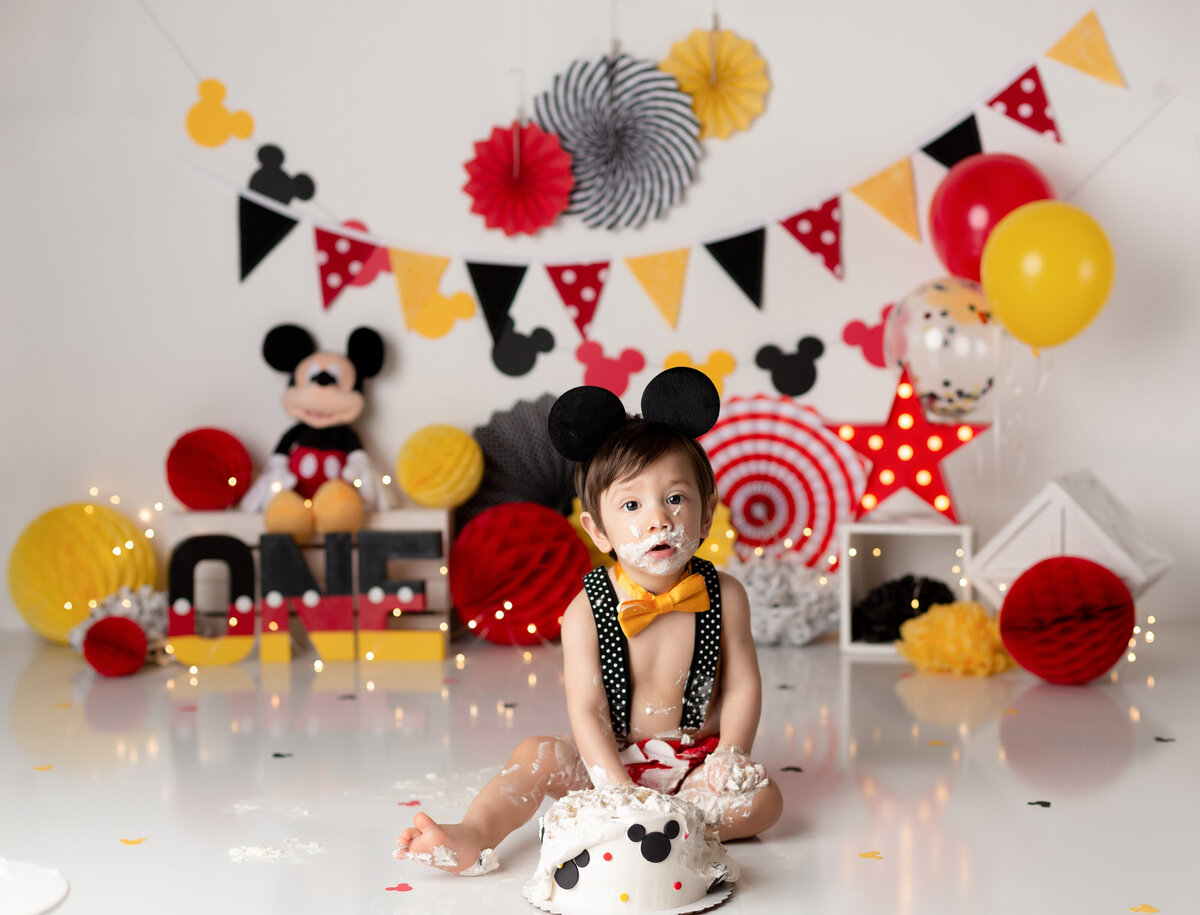 Mickey Mouse themed cake smash. Baby boy wearing red shorts and black and white polkadot suspenders and mickey mouse ears. Baby is smashing a mickey mouse decorated cake, with icing on his legs, arms, and face. In the background, there is red, yellow, and black decor.
