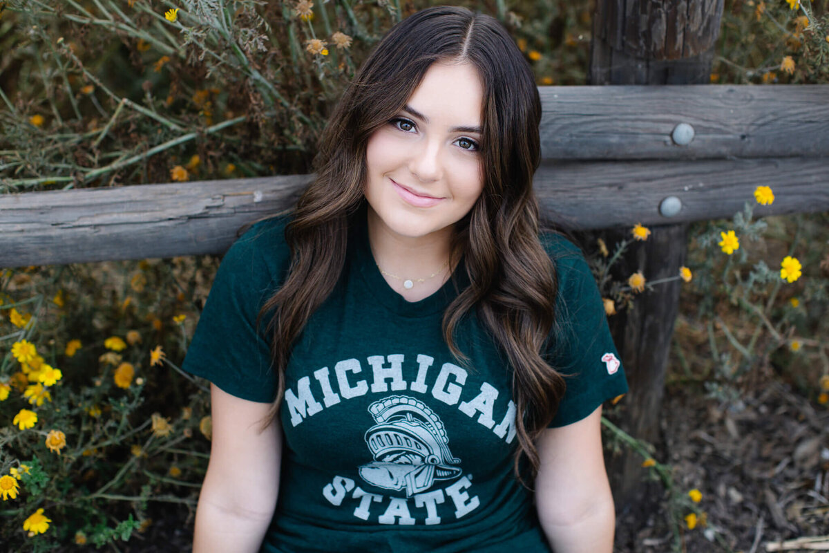 A young brunette woman wearing a green Michigan State T-shirt, sitting in front of a wooden park fence surrounded by yellow flowers, looking up at the camera.