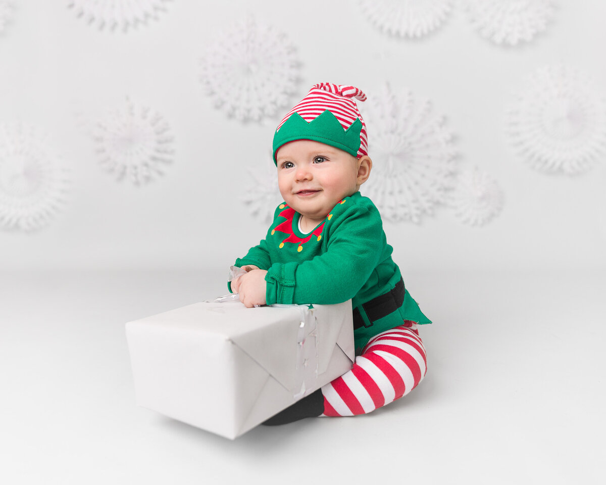 Image of a baby in an elf outfit holding a present on a Christmas snowflake backdrop in the studio. Taken by Norwich portrait photographer Claire Howes. Christmas Mini Sessions Norwich.