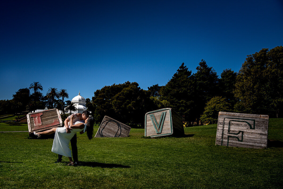 This playful and romantic engagement photo was captured at the Conservatory of Flowers in San Francisco. The couple is beautifully framed in front of large wooden letters spelling "LOVE," with the iconic white structure of the conservatory in the background. The bright blue sky and lush green lawn add to the joyful atmosphere of the shot, while the couple’s intimate dip captures their love and excitement. This location made for a perfect setting, blending both romance and fun into one memorable image.