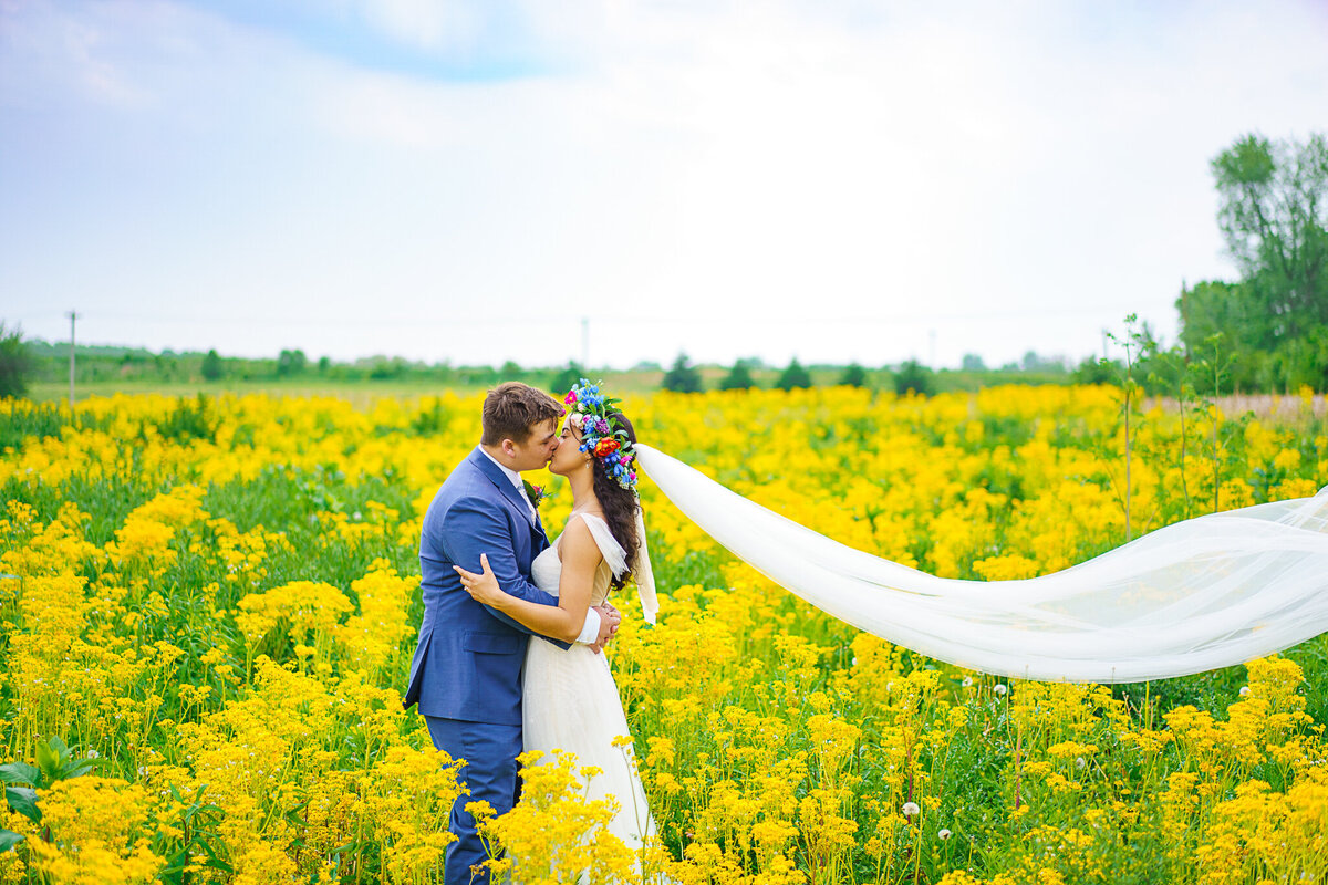 Bride and groom, Ronicia and Jack Henderson share a kiss in a field of yellow flowers as Ronicia's cathedral veil flows elegantly behind her.