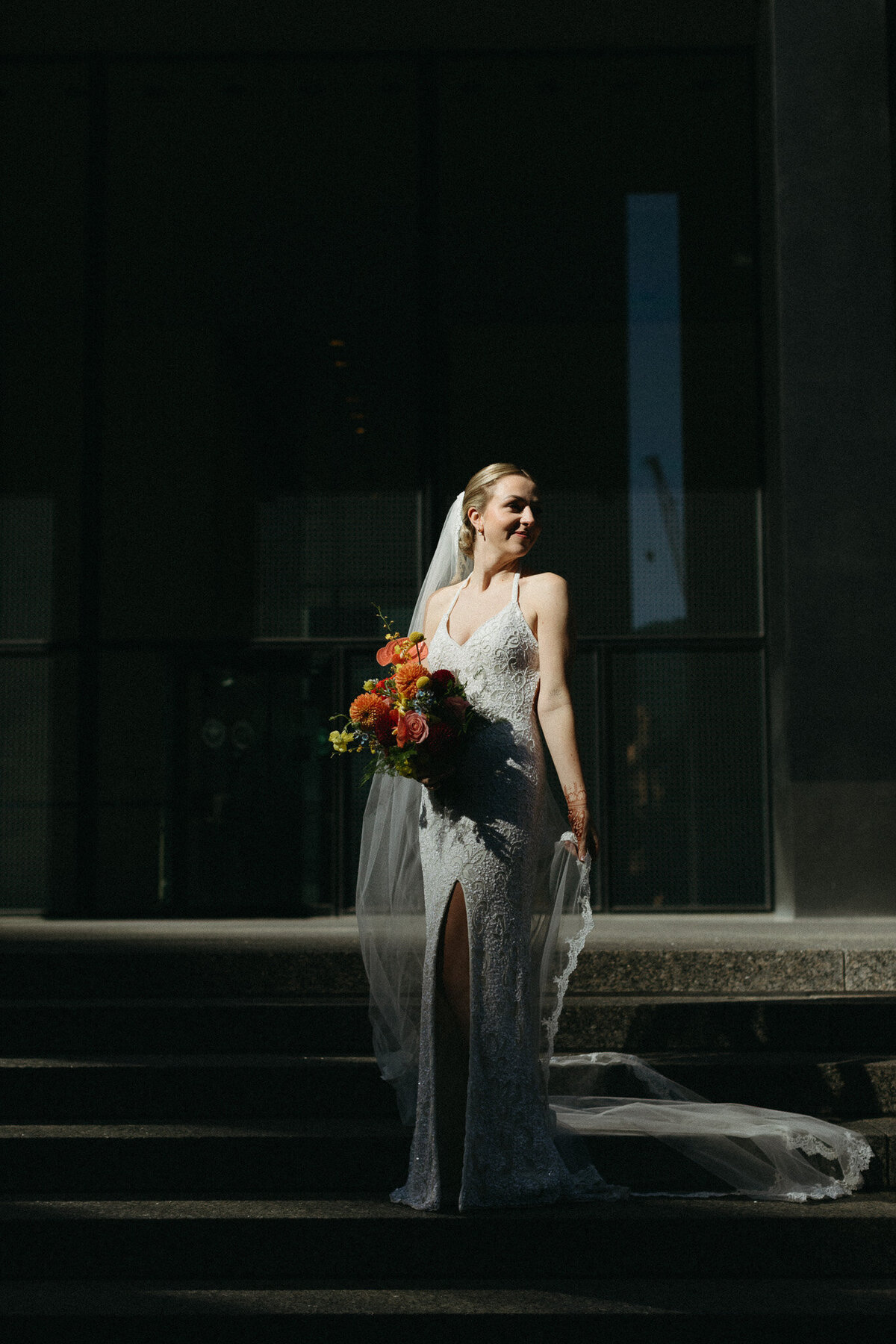 In the shadowed background, a bride in a white dress with a slit holds vibrant orange flowers on the steps, captured beautifully by Ontario destination wedding photographer, Jennifer van Son at Steam Whistle Brewing.