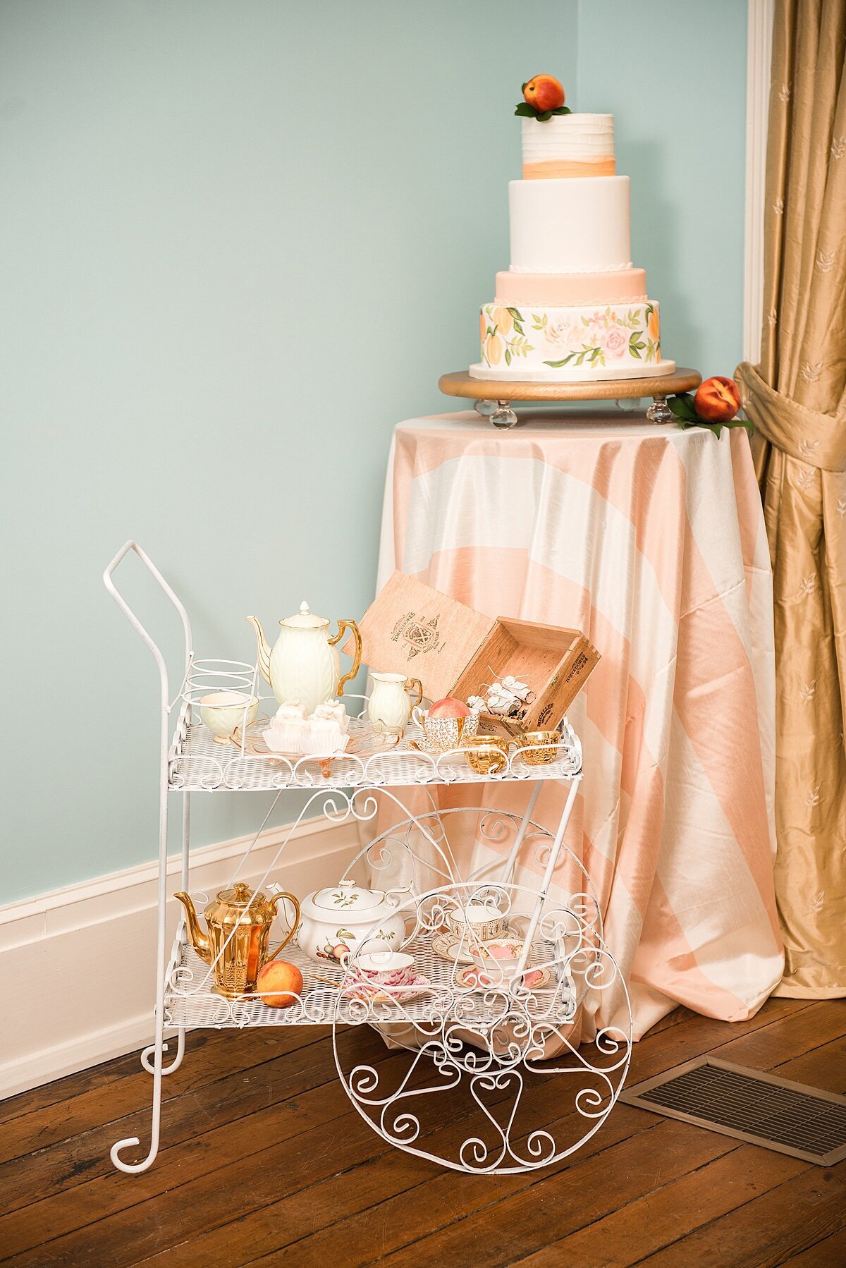 In the light blue front parlor of Ravenswood Mansion, the white wire tea cart has a full display of vintage china tea cups and vintage china tea pots as well as a vintage copper coffee pot and a selection of loose leaf teas. Behind the tea cart, is a tall cocktail table with a peach, raw silk table cloth topped with a five tier hand painted peach blossom wedding cake with a fresh peach cake topper and peach accents.