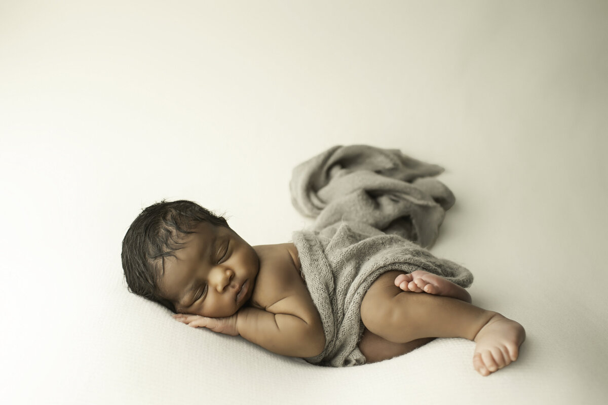 Newborn baby boy peacefully sleeping on a soft white backdrop, gently wrapped in a cozy grey fabric. His tiny hands peek out as he rests comfortably, with a serene expression and delicate features highlighted by soft, natural lighting.