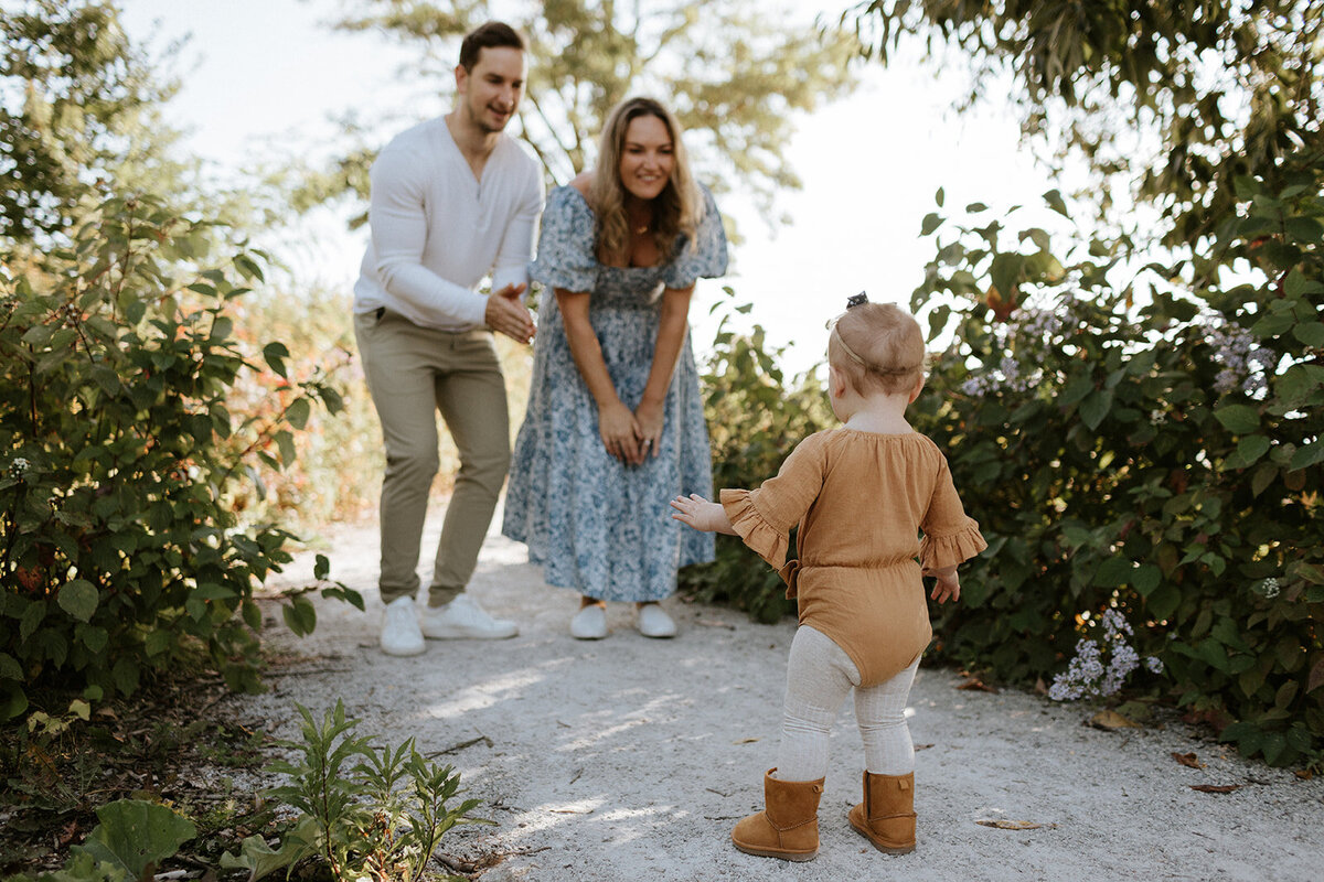 A toddler in a brown outfit walks toward a man and woman smiling and clapping on a green, scenic path, beautifully captured by an Oakville family photographer.