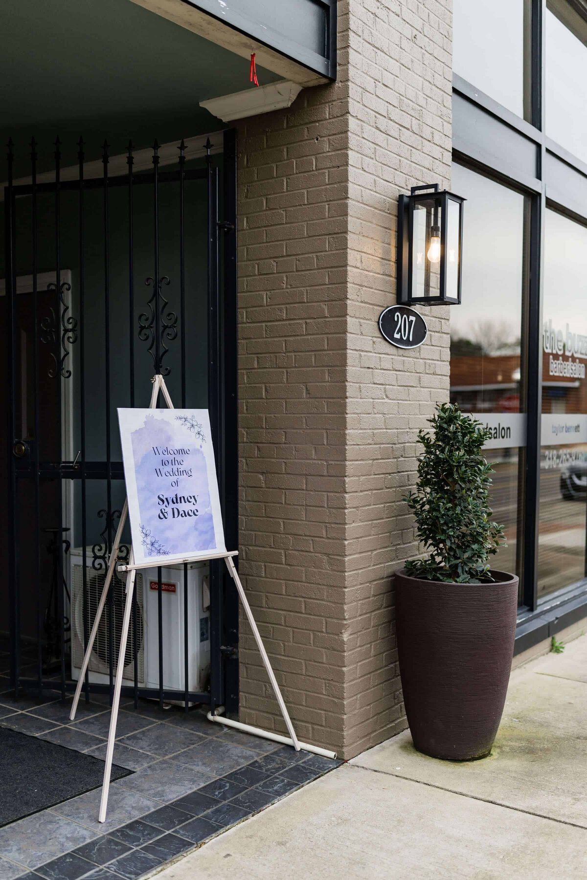 A sign on an easel near a building entrance reads Welcome to the wedding of Sydney and Dace. A potted plant and a lantern-style light fixture are beside the door, with the address number 207 on the wall.