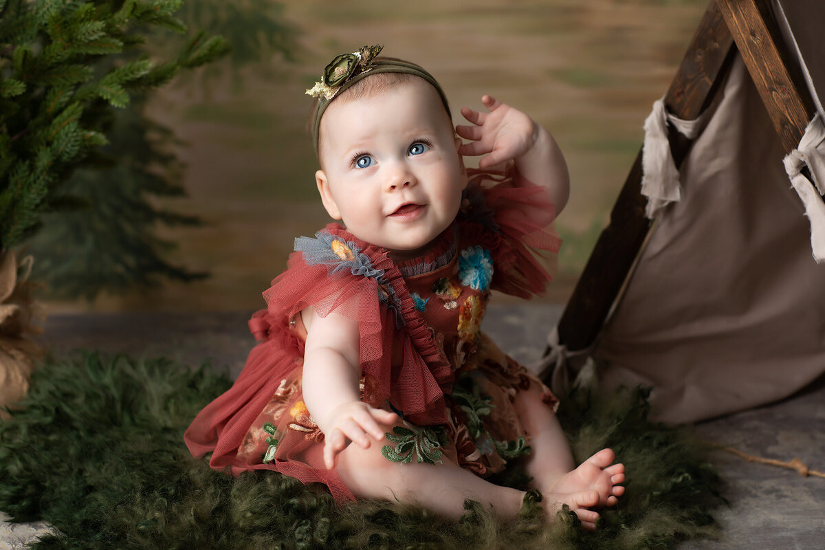 A bright-eyed baby girl in a rust-colored embroidered dress sits on a soft green rug beside a tiny canvas tent, with forest elements surrounding her. Captured by a Dallas baby milestone photographer, this whimsical outdoor-inspired scene beautifully celebrates a joyful milestone moment.