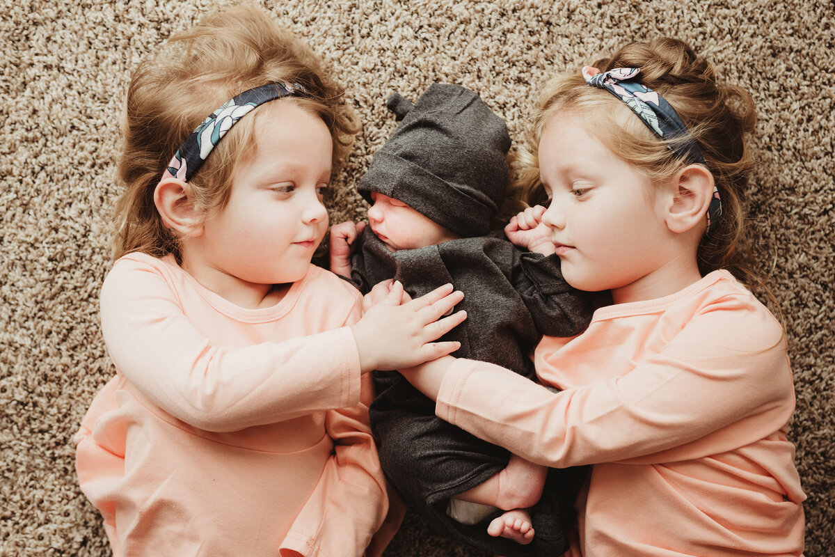 Baby sisters are peacefully lying with their newborn brother on the floor.