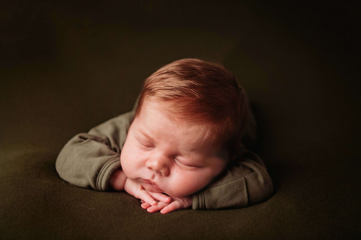 A red headed newborn baby boy, sleeping peacefully in green pajamas on a matching green blanket. He has his hands tucked gently under his chubby cheeks, as he sleeps on his belly inthis gorgeous newborn photo by a Mobile, AL newborn photographer