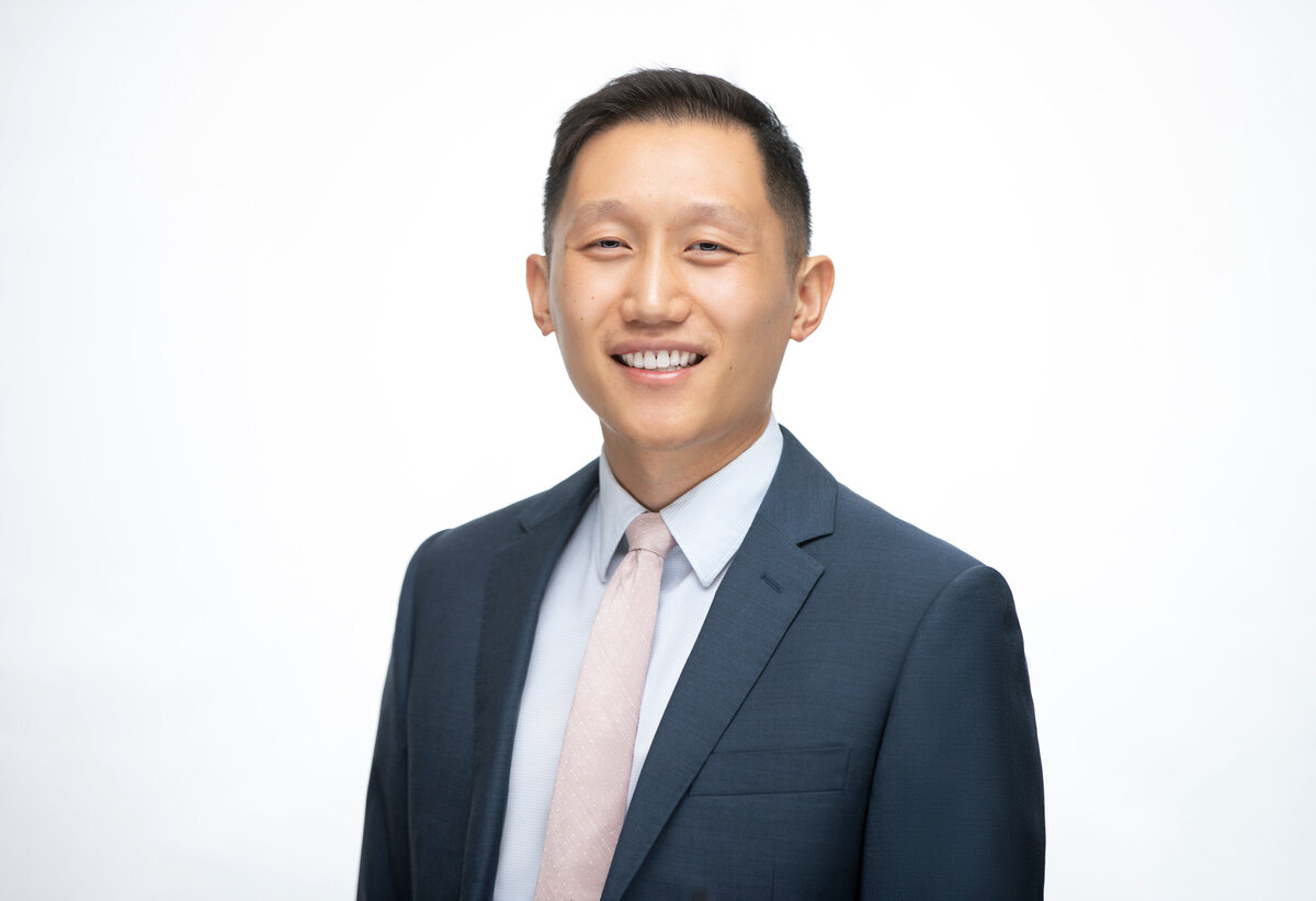 An Asian American professional doctor in a blue suit jacket poses for a professional headshot on a white background for Janel Lee Photography studios in Cincinnati Ohio