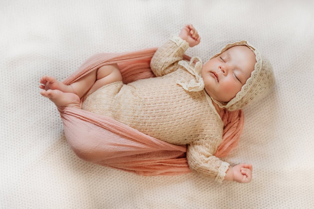 newborn girl in tan onesie and bonnet with arms up sleeping