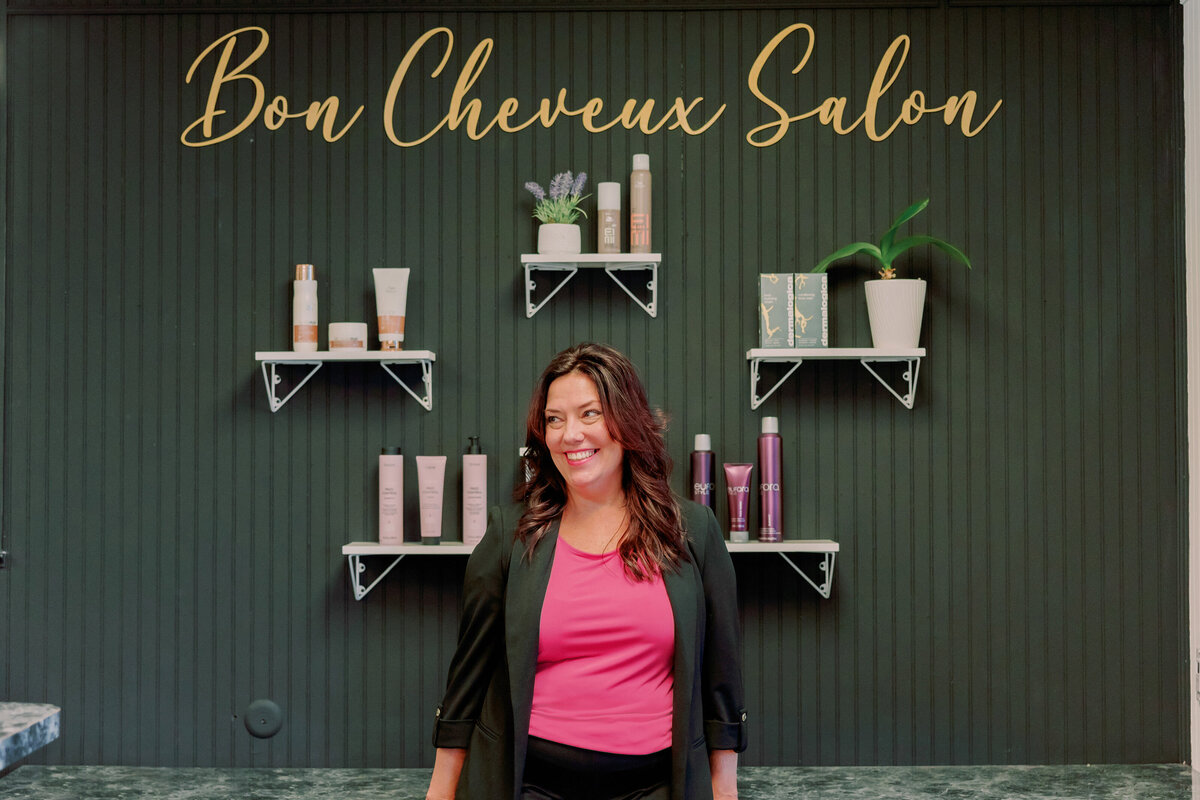 A hairstylist poses in front of a wall with products that says "Bon Cheveux Salon."