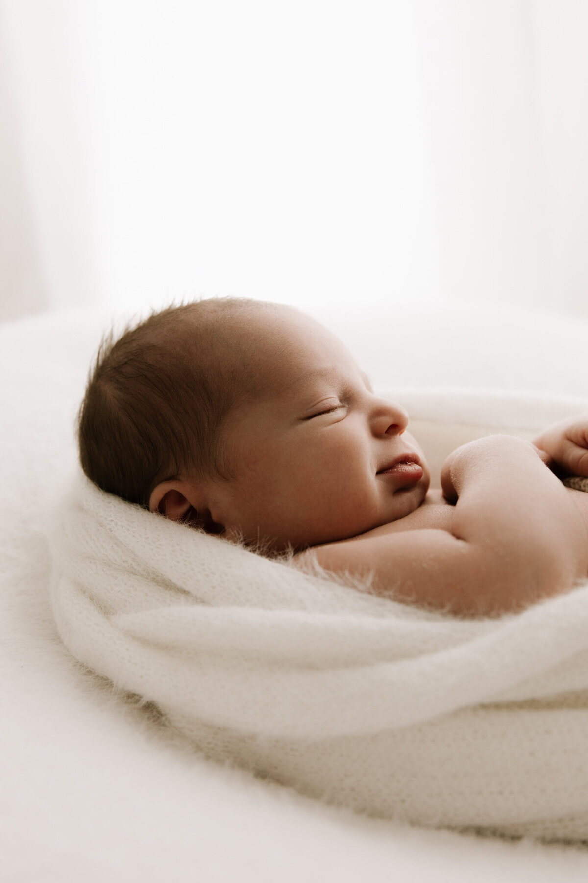 A close-up profile of a newborn baby peacefully smiling in their sleep, swaddled in a soft white wrap. The baby lies on a cozy white surface, bathed in gentle, diffused studio light. A serene and heartwarming moment captured during a newborn photography session.