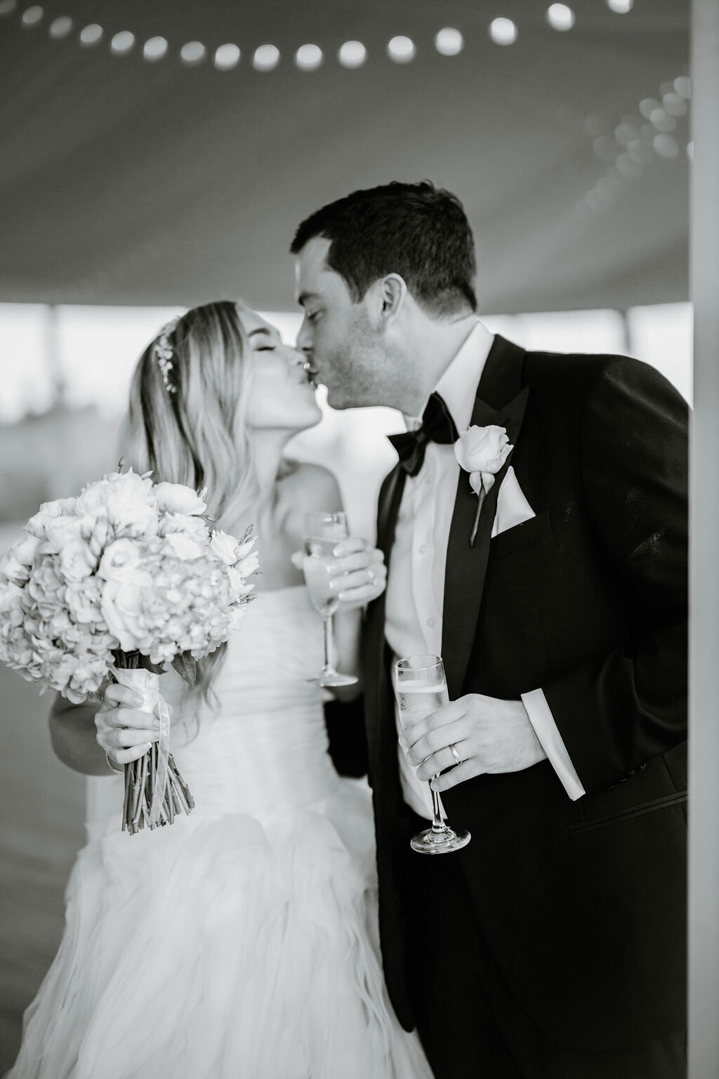 Rhode Island Wedding Photographers | A couple kisses on their wedding day, holding champagne and a bouquet. The groom wears a tuxedo, the bride a strapless wedding dress. 