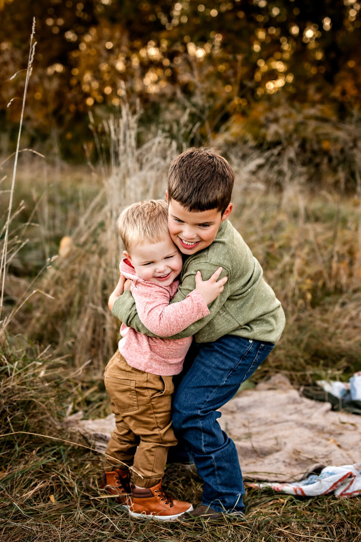Two young children joyfully embracing each other in a grassy field, perfect for capturing outdoor portraits. One child dons a pink sweater with tan pants, while the other sports a green sweater and jeans. Their broad smiles reveal the playful moment they're enjoying in nature's embrace.