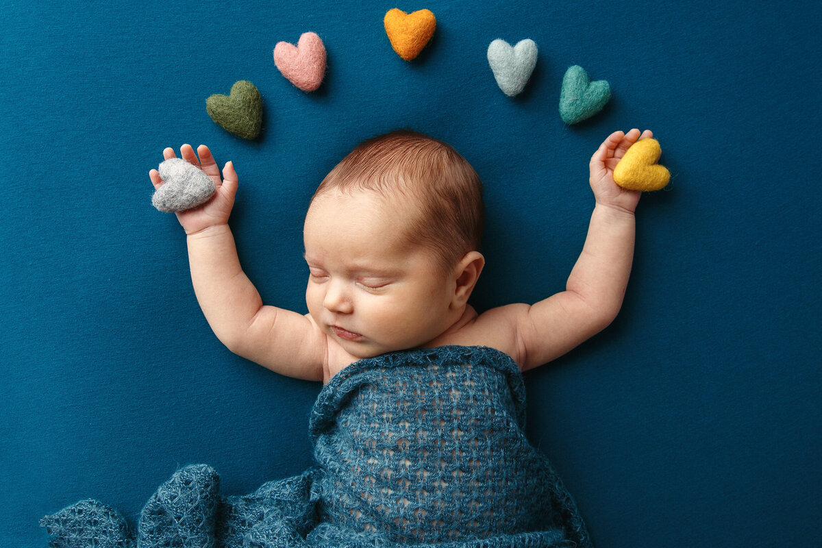 Newborn baby asleep on his back photographed on a blue blanket with his hands reaching up to an arch of colored hearts