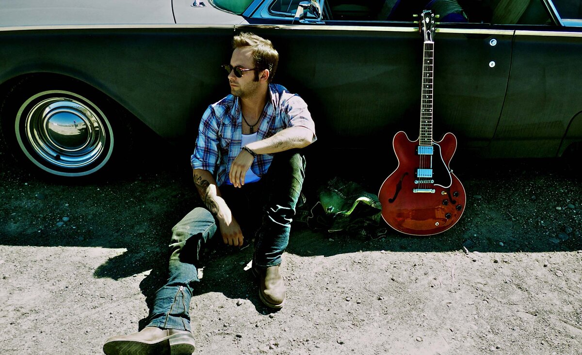 Male musician portrait Dallas Smith wearing plaid shirt blue jeans sitting against vintage black car red guitar leaning beside