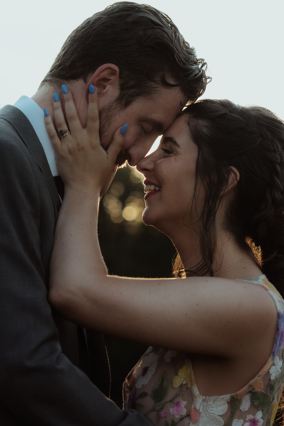 A bride and groom touch foreheads and noses, the bride touching his face, as they laugh candidly in an intimate close-up backlit portrait.