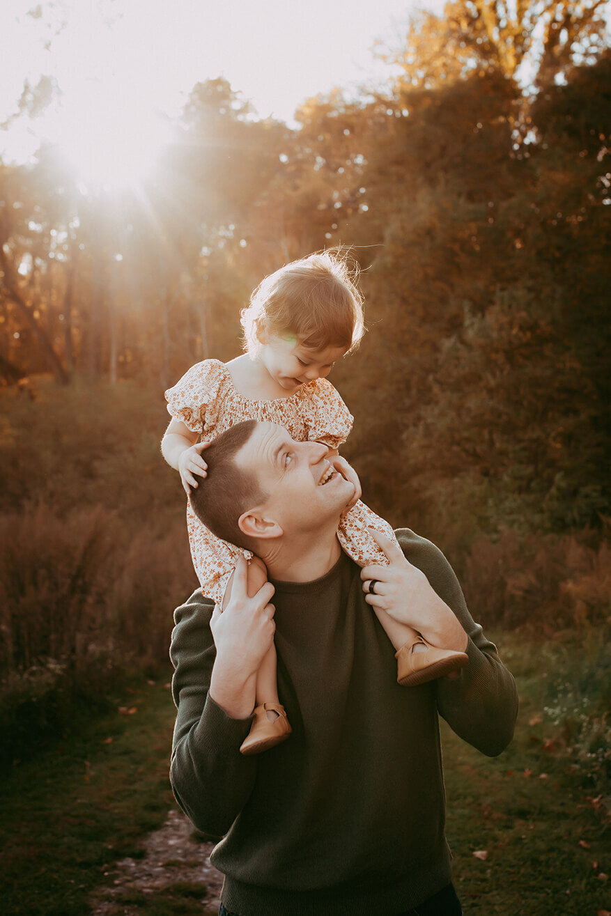 a little girl sitting on her dads shoulders looking at each other during golden hour