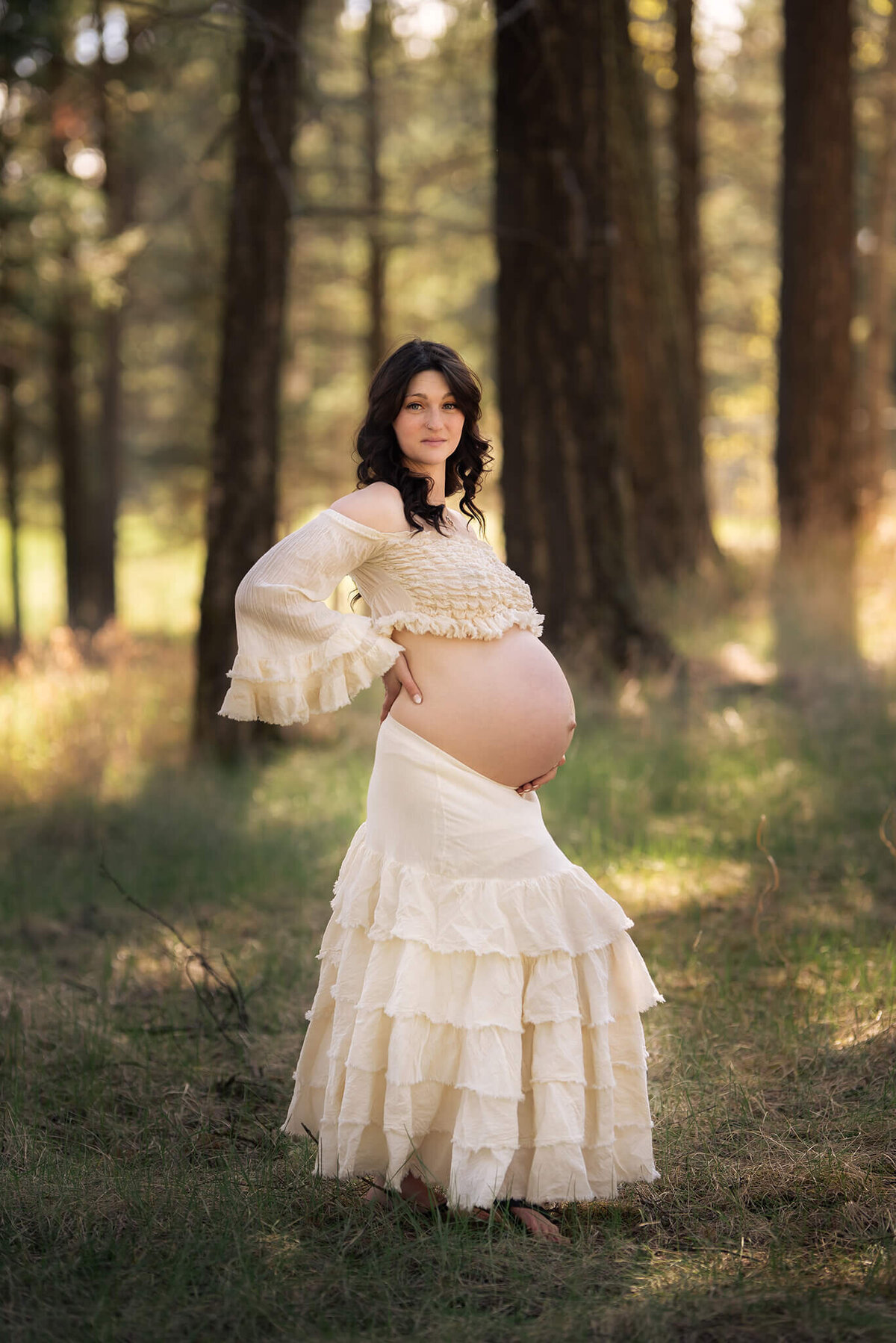 A pregnant woman with brown curly hair stands in the forest in a matching cream coloured top and skirt. Taken by Brittany Danielle