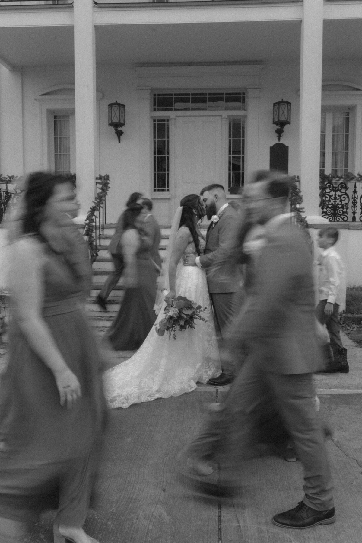 A black and white photo of a wedding couple kissing while their wedding party is caught in motion blur while walking. 