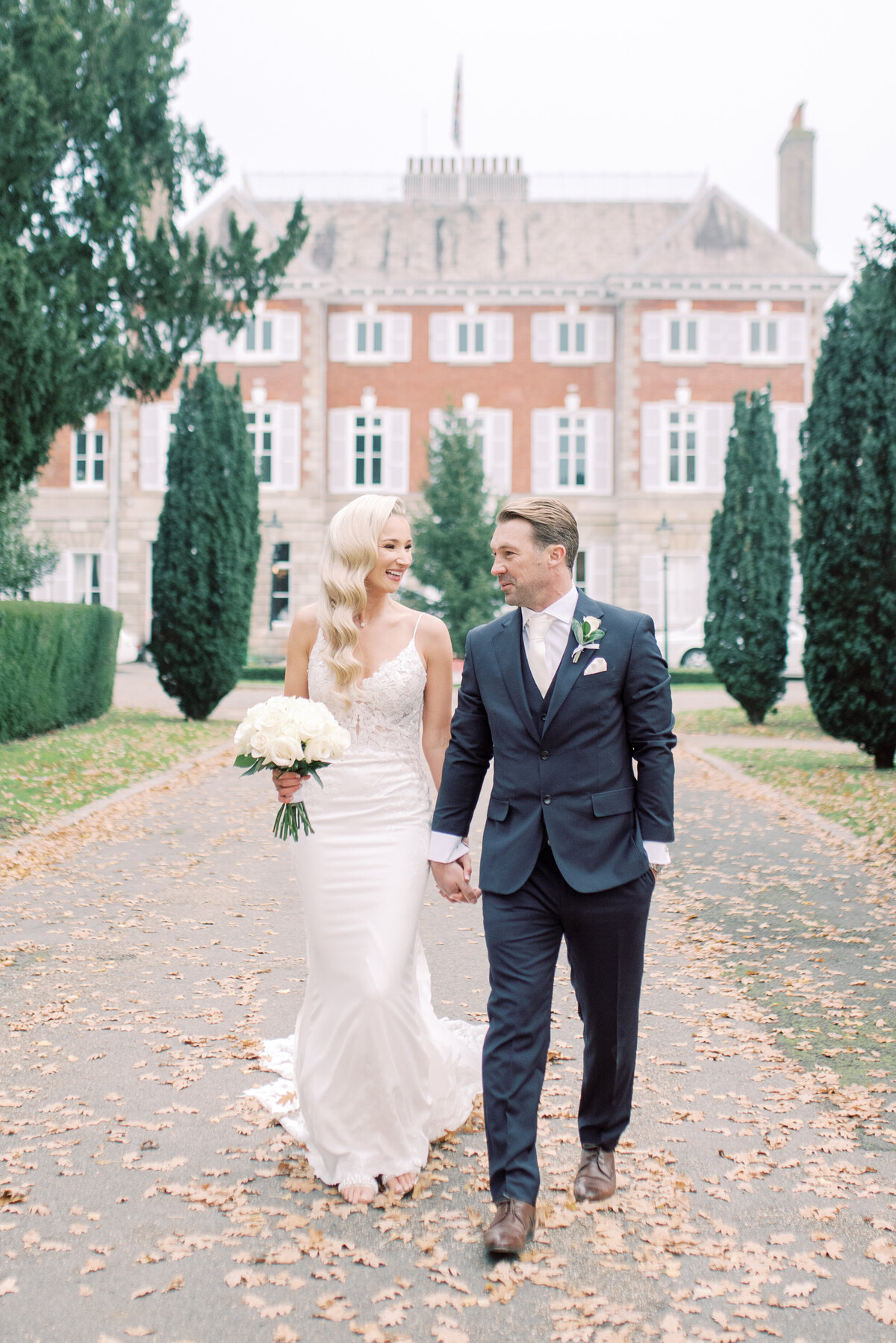 Light and airy style image bride and groom walking toward sthe camera holding hands, looking at each other and smiling. The bride is wearing a fitted dress with lace detailing and the groom is in a dark blue suit